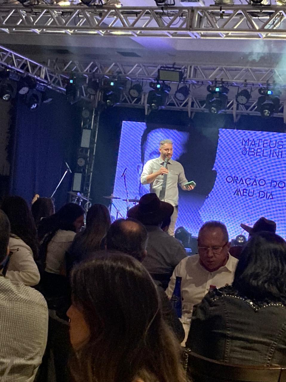 Pedro Prudente no palco durante um evento corporativo à noite, microfone em mãos e a plateia sentada às mesas em primeiro plano